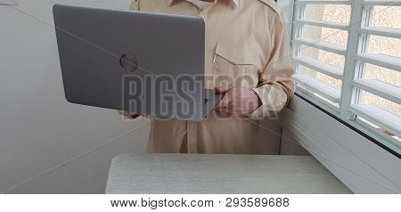 A Man Wearing Light Colored Official Shirt Stands In The Corner Near Window Holds In Hands Open Lapt