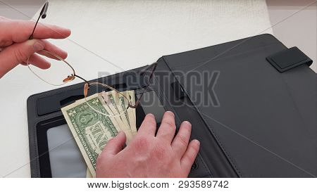 Eyeglasses In Male Left Hand While His Right Hand Lays On American Dollars Banknotes In Black Open P