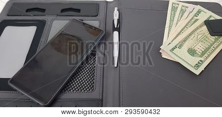 American Dollar Bills Catched In A Magnet Pocket Of Black Leather Folder With A Pen And A Smart Phon