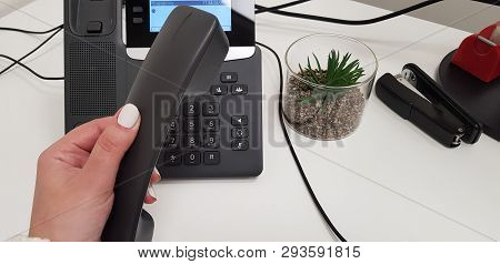 Woman Wearing White Nail Polish Holds Black Handset In Left Hand Over Office Phone Near A Stapler An