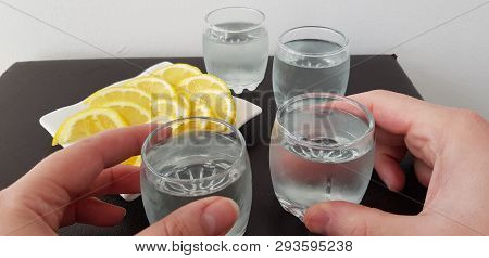 Two Hands Hold Glasses With Vodka Over Brown Tablewith White Plate With Lemon Slices And Two More Gl