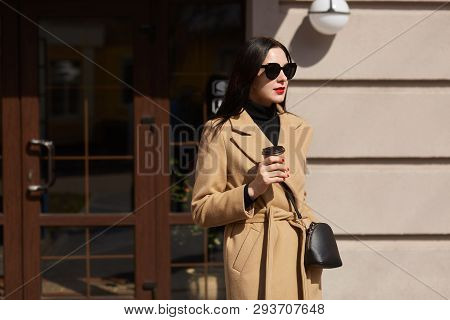 Beautiful Brunette Young Woman Holds Cup Of Coffee In Her Hands, Walks Along Sunlighted Streets, Wea
