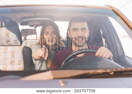 Astonished Young Woman Sits On Back Seat In Car, Indicates At Windscreen With Interested Expression 