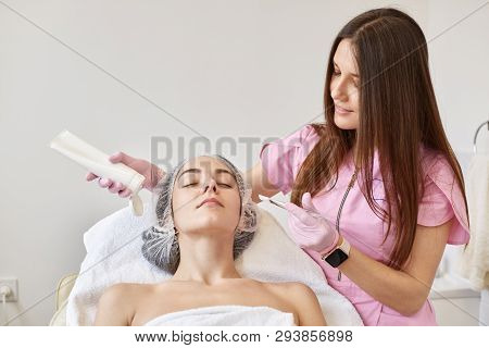 Cosmetologist Applying Facial Mask On Beautiful Woman Skin. Attractive Woman Enjoying Face Mask Trea