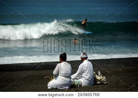 An elderly Balinese couple getting ready for morning ritual while two surfers do their morning ritual as well.