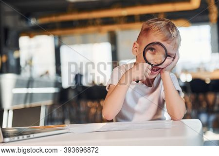 Smart Child In Casual Clothes With Laptop On Table Have Fun With Magnifying Glass.