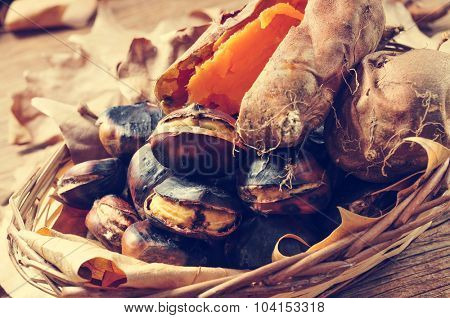 some roasted chestnuts and some roasted sweet potatoes in a wicker basket with autumn leaves, on a rustic wooden table