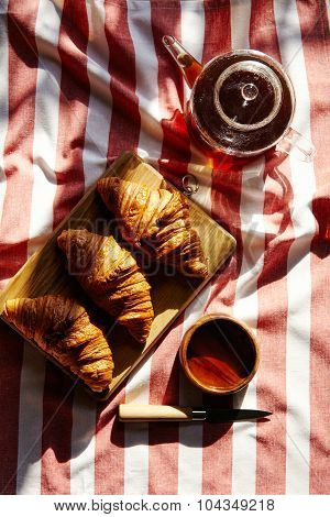 Three French Freshly Baked Croissants On Wooden Plate With Teapot And Knife On Striped Cloth