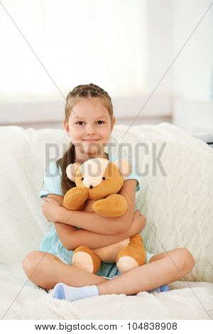 Beautiful little girl with teddy bear sitting on sofa, on home interior background