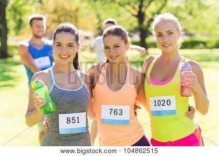 fitness, sport, friendship, marathon and healthy lifestyle concept - happy young sporty women with racing badge numbers and water bottles outdoors