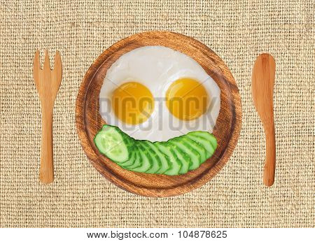 Fried Eggs And Fresh Cucumber On Wooden Plate, Fork And Knife On The Llinen Texture Background