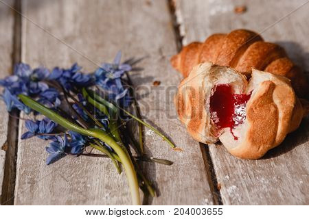 Delicious croissant on wooden background. Sweet breakfast with fruit jam and spring blue snowdrops nearby. Morning fresh and tasty meal. Food photography close up