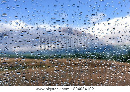 Colorful view of the mountains with fog forest blue sky and clouds through the window glass of the car covered by rain drops