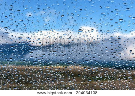 Colorful view of the mountains with fog forest blue sky and clouds through the window glass of the car covered by rain drops