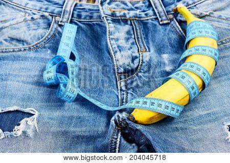 Banana wrapped with blue measure tape on jeans selective focus. Mens denim pants crotch with banana imitating male genitals. Jeans zipper and pocket close up. Health and male sexuality concept.