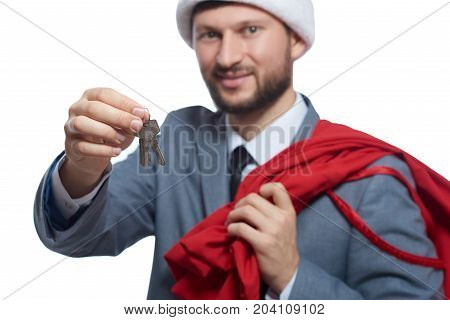 Portrait of man in gray suite, red cap like santa claus, holding full bag over shoulder, holding, giving key from house. Santa smiling at camera giving luxury present. Christmas, new year concept.
