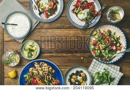 Flat-lay of healthy dinner table setting. Fresh salad, grilled vegetables with yogurt sauce, pickled olives over wooden background, copy space, selective focus. Clean eating, vegan, vegetarian food