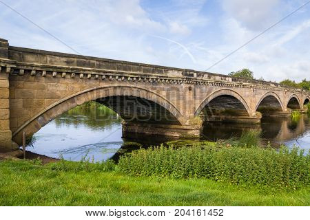 Stone Bridge over The River Trent between Repton and Willington Derbyshire. Showing the river, cloudy sunny blue sky, green folage foreground, reflection of bridge in water.
