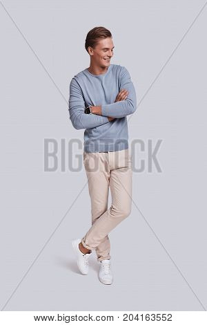 Young and confidence. Full length of handsome young man keeping arms crossed and smiling while standing against grey background