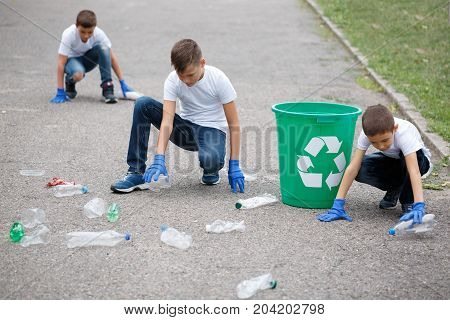 Photo of charity environment concept. Sunny day. Children in blue jeans and white t-shirt with colorful: blue and green recycle bin with label. Cleaning in park toghether. Team work.