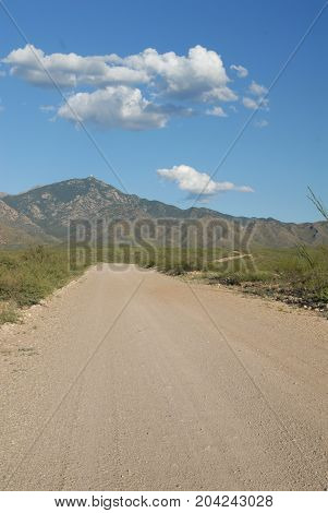 A gravel and dirt path leading into the mountains in southern Arizona with a blue sky and puffy white clouds.