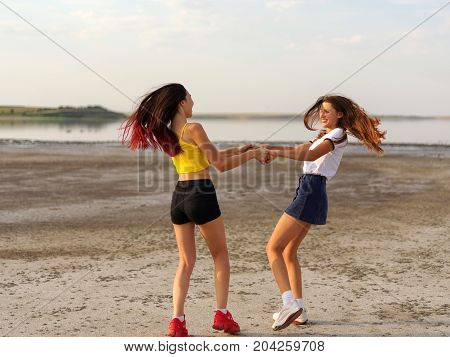 Two girlfriend reel round circling in dance in the summer daisy. Summer field against blue sky with white clouds. Woman play outdoor in short stylish clothes. Friendship concept - happy young women.