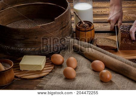 The hand cuts black bread on the background of ingredients for cooking bread. eggs honey milk butter flour sieve rocking chair for dough on a background of burlap and wooden background