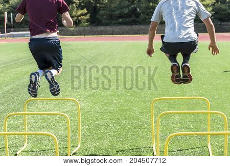 Two high school athletes bound over yellow hurdles on a green turf field during afternoon practice.