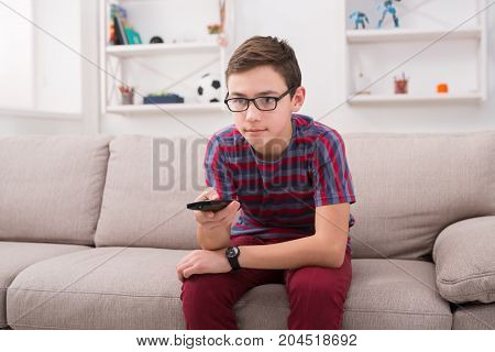 Teenager boy watching television, using TV remote control while sitting on couch in living room at home