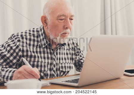Mature man using laptop and writing in notepad at home desk. Education, recipe concept