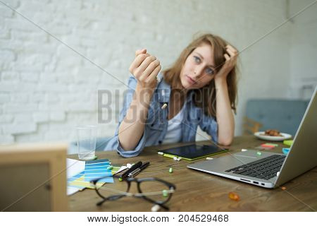 Unhappy overworked female photographer sitting at desk with laptop pc and graphic tablet spilling out colorful pills feeling exhausted spending sleepless night retouching images. Selective focus