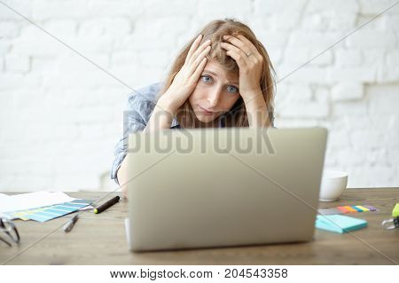 Picture of overworked beautiful freckled young woman freelancer holding hands on her head and looking at screen of laptop pc in frustration feeling tired and stressed out because of deadline