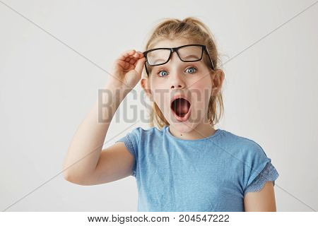 Charming little blonde lady with blue eyes reises glasses with hand in happy excitement of new big bed in her sleeping room as a present