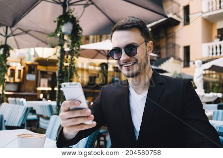 Professional and successful businessman is sitting at the table in restaurant. He has his glasses on and looking to the phone's screen. Apparently he has a Skype call or checking his social media's profile because he is smiling. Close up. Cut view