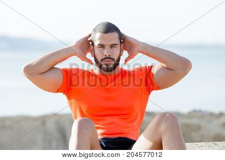 Closeup portrait of serious young man wearing sportswear, looking at camera, keeping hands behind head and doing sit-ups outdoors in summer. Front view.