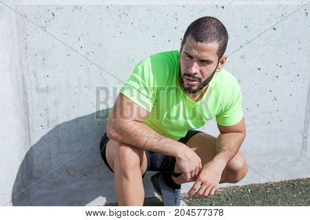 Closeup portrait of tired young handsome strong man wearing sportswear, looking away and sitting on hunkers at concrete wall