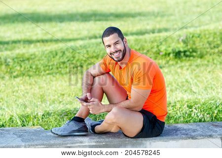 Cheerful muscled guy smiling at camera on stroll and sitting on concrete slab. Happy modern sportsman with smartphone spending time in park after training. Technology and sport concept