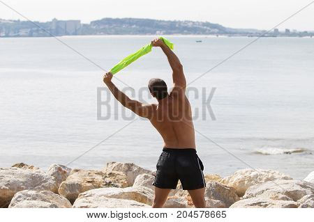 Closeup of shirtless young handsome strong man holding t-shirt, stretching body and doing side bend exercise with sea in background. Back view.