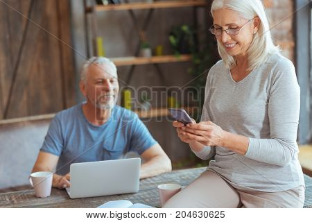 In love with technologies. Positive beautiful elderly woman using her smart phone and sitting on the table while her husband working on the laptop