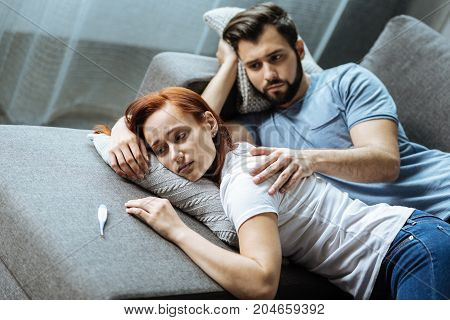 You need rest. Sad cheerless bearded man looking at his ill girlfriend and holding her shoulder while showing his care