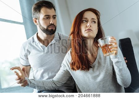 Do not touch me. Unhappy sad young woman holding a glass of whisky and not allowing her boyfriend to take it away while being alcohol addicted