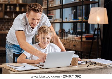 Careful selection. Joyful young man standing behind his pre-teen son and pointing at the laptop, helping the boy to choose necessary pieces of information for his school project