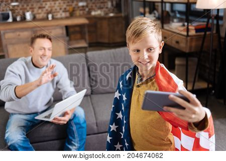 Happy family. Cheerful pre-teen boy wearing a US flag on his shoulders and taking a selfie with his father sitting in the background, waving at the camera while working on laptop
