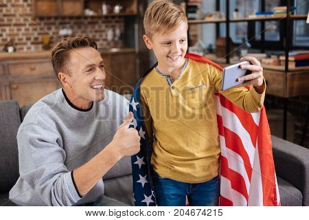 Patriotic family. Charming pre-teen boy wearing a US flag on his shoulders and taking a selfie with his father showing a thumbs up