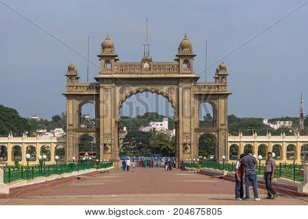 Mysore India - October 27 2013: Closeup of brown stone main East Gate to Mysore Palace under blue sky. Yellow wing corridor on both sides. Visitors and city view with trees.
