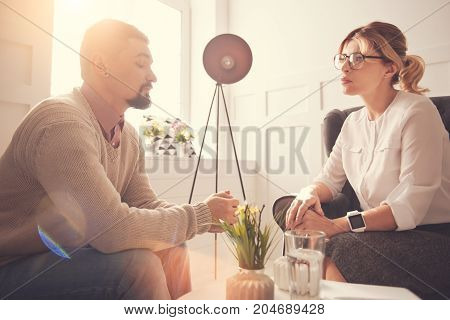 Working with patients. Nice serious good looking therapist sitting in the armchair and listening to her patient while having a psychological session with him