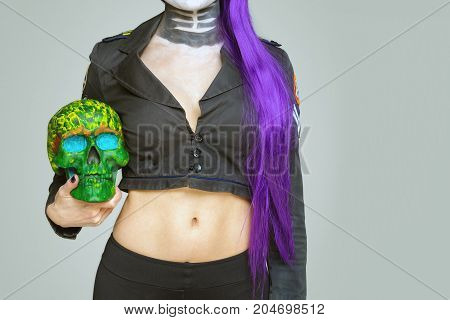 girl with makeup and purple hair holds a painted skull in her hand on a gray background. Mexican celebration of the Day of the Dead Dia de los Muertos and the Roman Catholic holiday All Souls Day