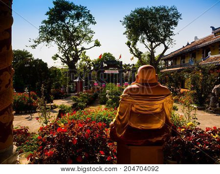 HOIAN, VIETNAM, SEPTEMBER, 04 2017: Back view of a budha in an ancient temple with a beautiful jarden with colorful flowers at hoian, in a sunny day in Vietnam.