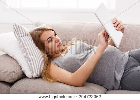 Young pregnant woman using digital tablet. Expectant blonde texting with tablet sitting on the floor. Networking, modern technologies, pregnancy, communication concept