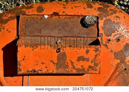 A lone wasp nest hangs from the lid of an old tool box mounted on an orange tractor fender.